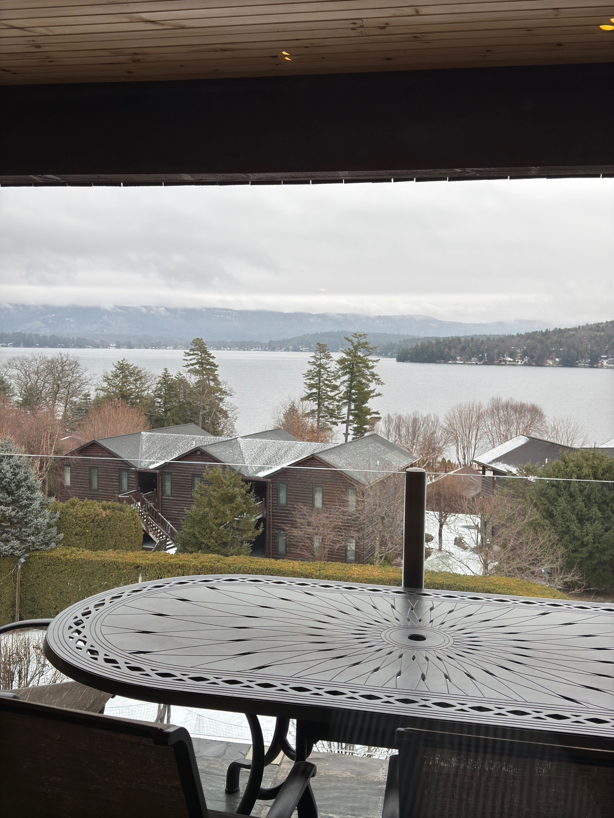A view from a covered outdoor area with a metal table and chairs, overlooking a lake and houses in the distance.