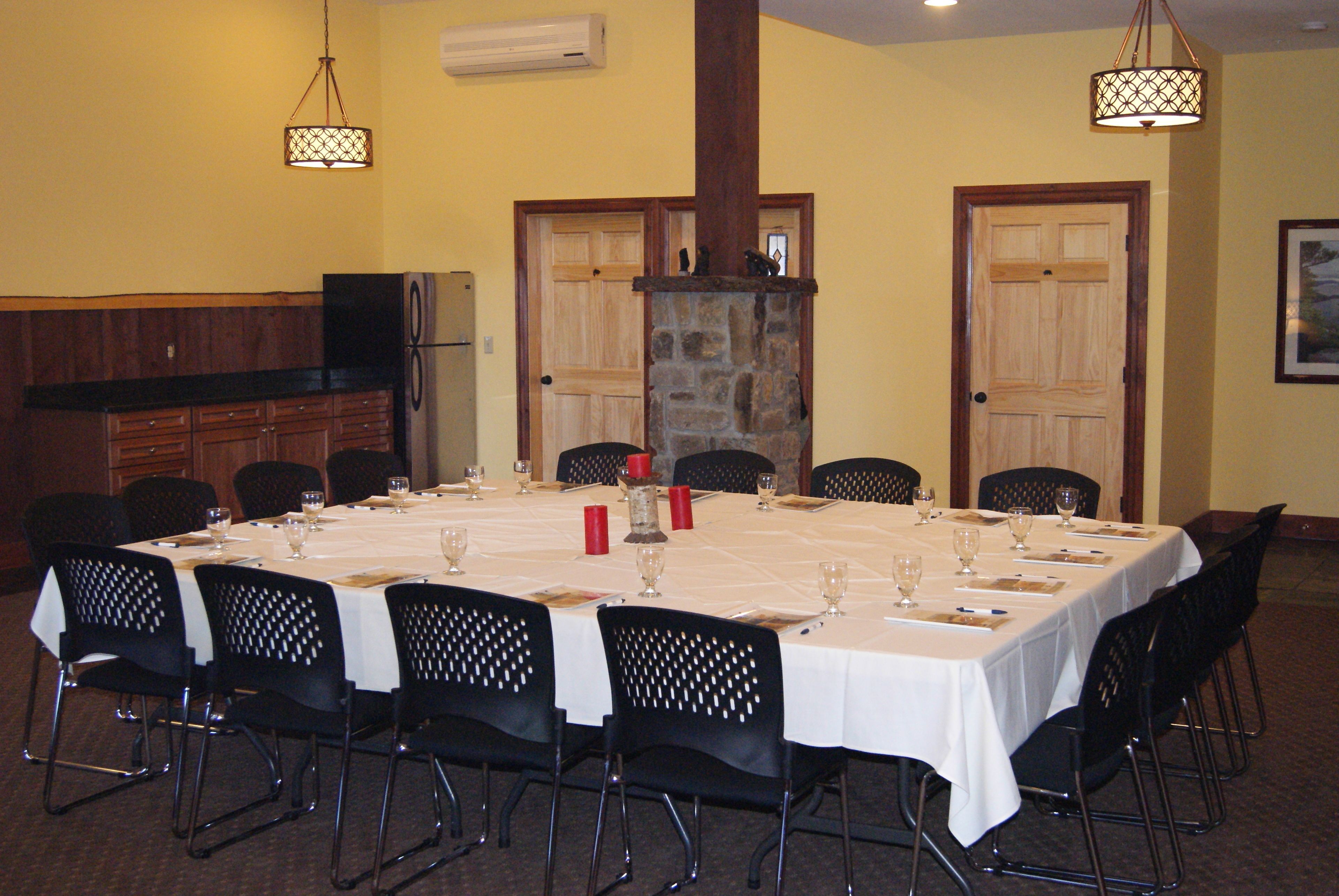 A dining area with a long table set for a meal, surrounded by chairs. There is a stone fireplace, wooden doors, and a kitchen area with a refrigerator and cabinets.