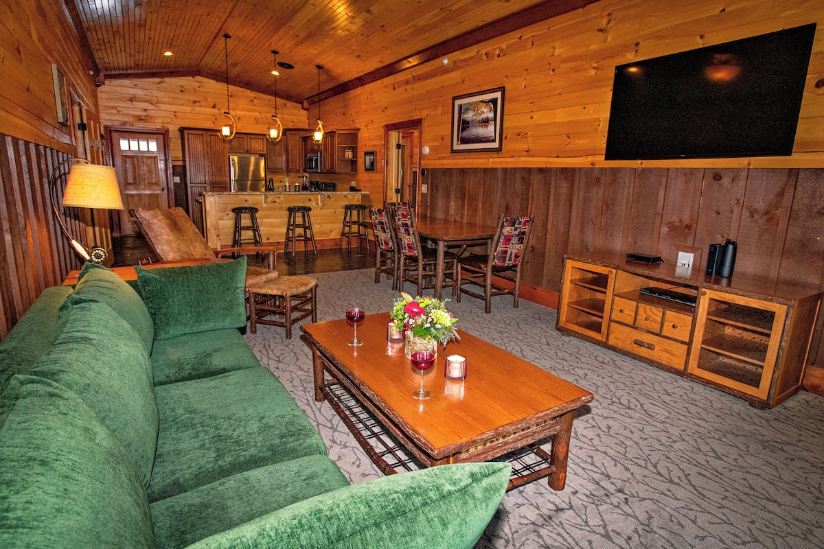 A cozy log cabin interior with a green sofa, wooden dining table, and a TV mounted on the wall.