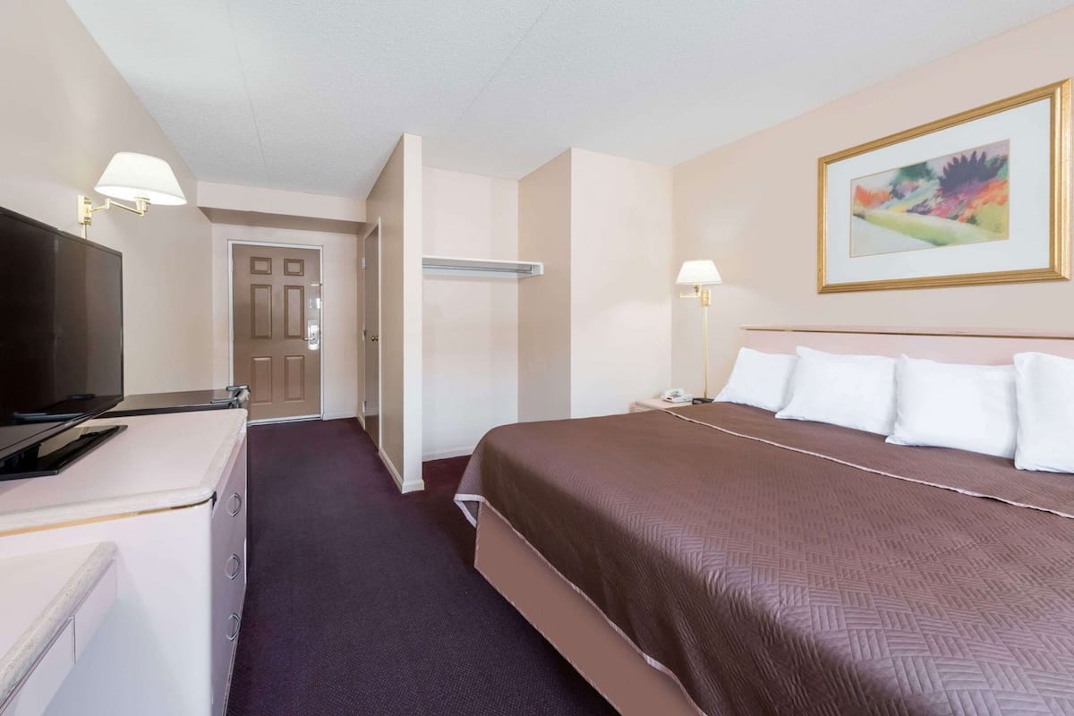 Una habitación de hotel con cama, lámparas de noche, televisor, armario y puerta que da a otra estancia.