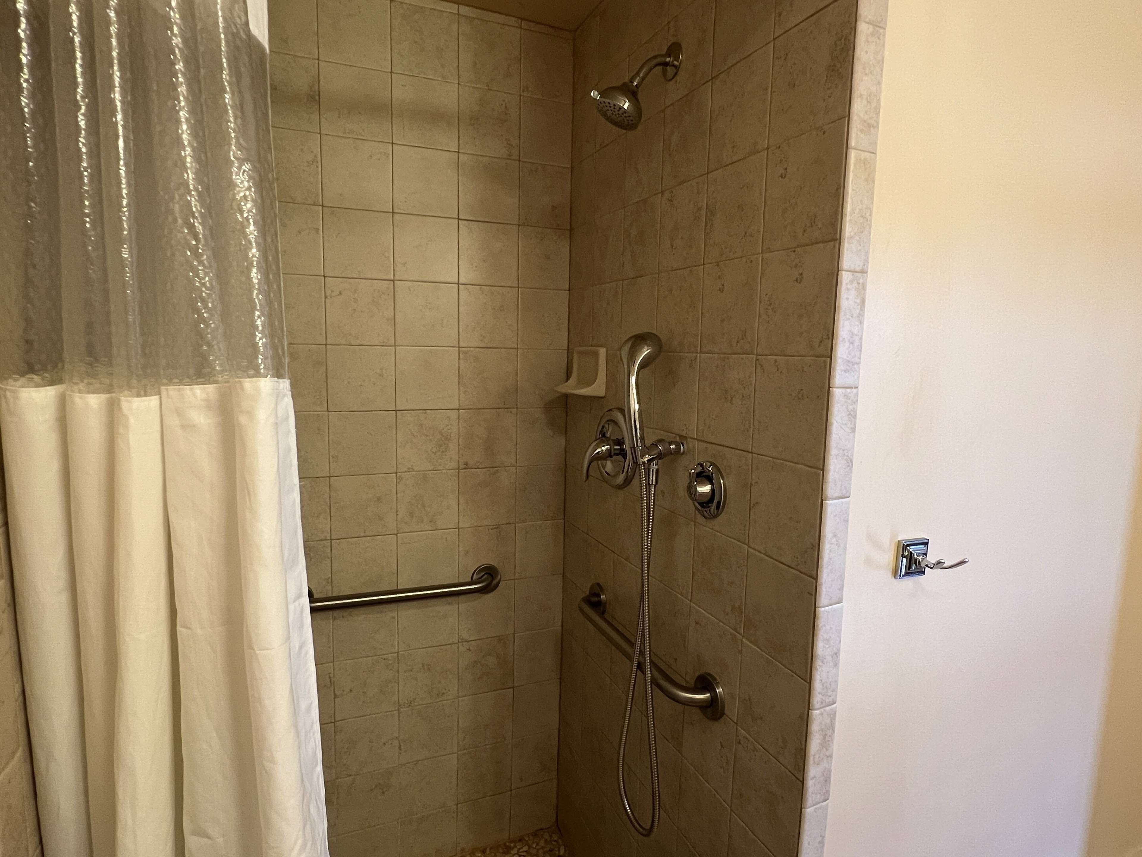 A shower area with tiled walls, a showerhead, a handheld showerhead, and a grab bar.