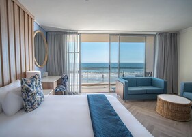 Quarto de hotel com cama, escrivaninha, poltrona, sofá e vista para o oceano.