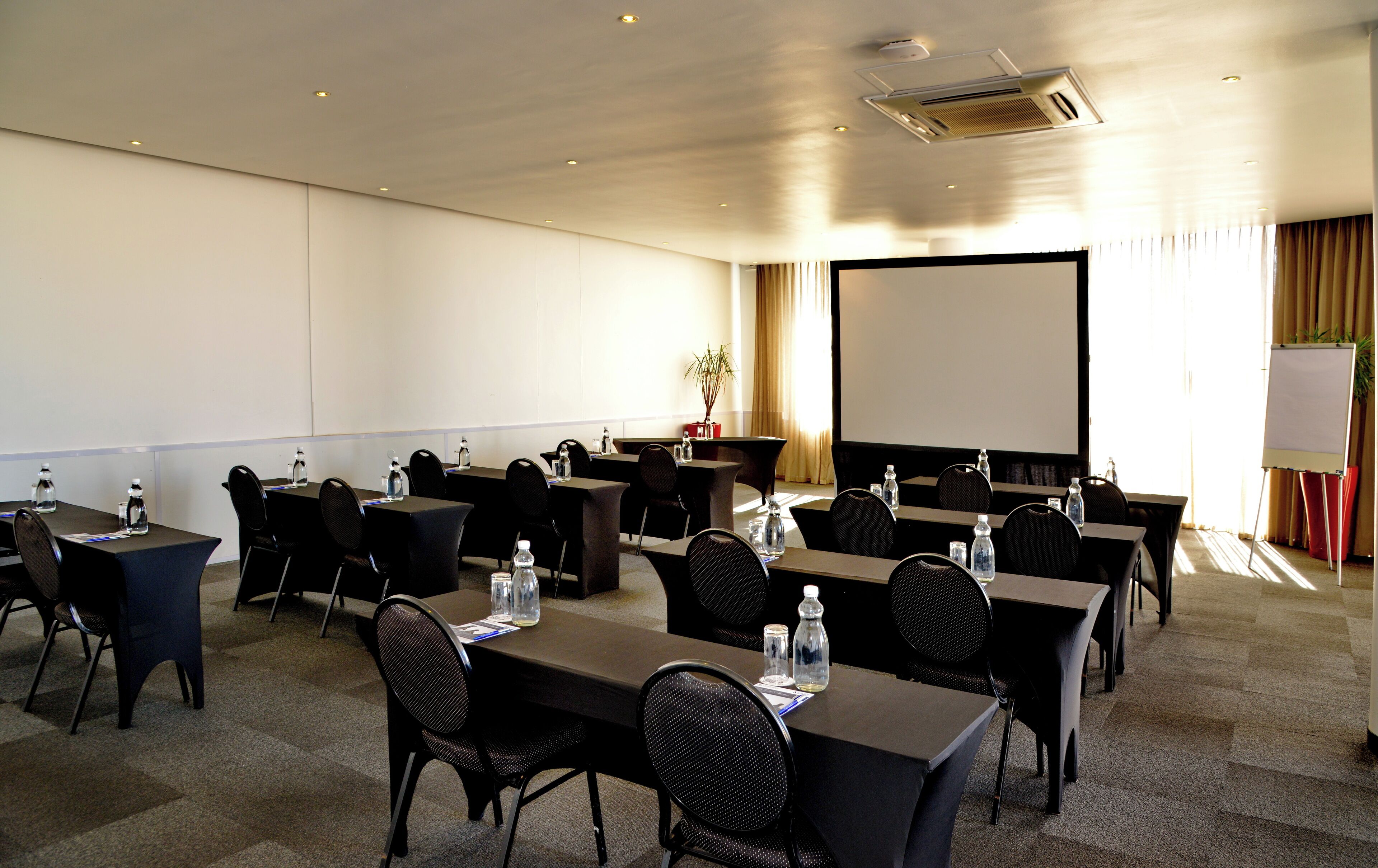 A conference room with multiple tables and chairs, equipped with water bottles and a projector screen.