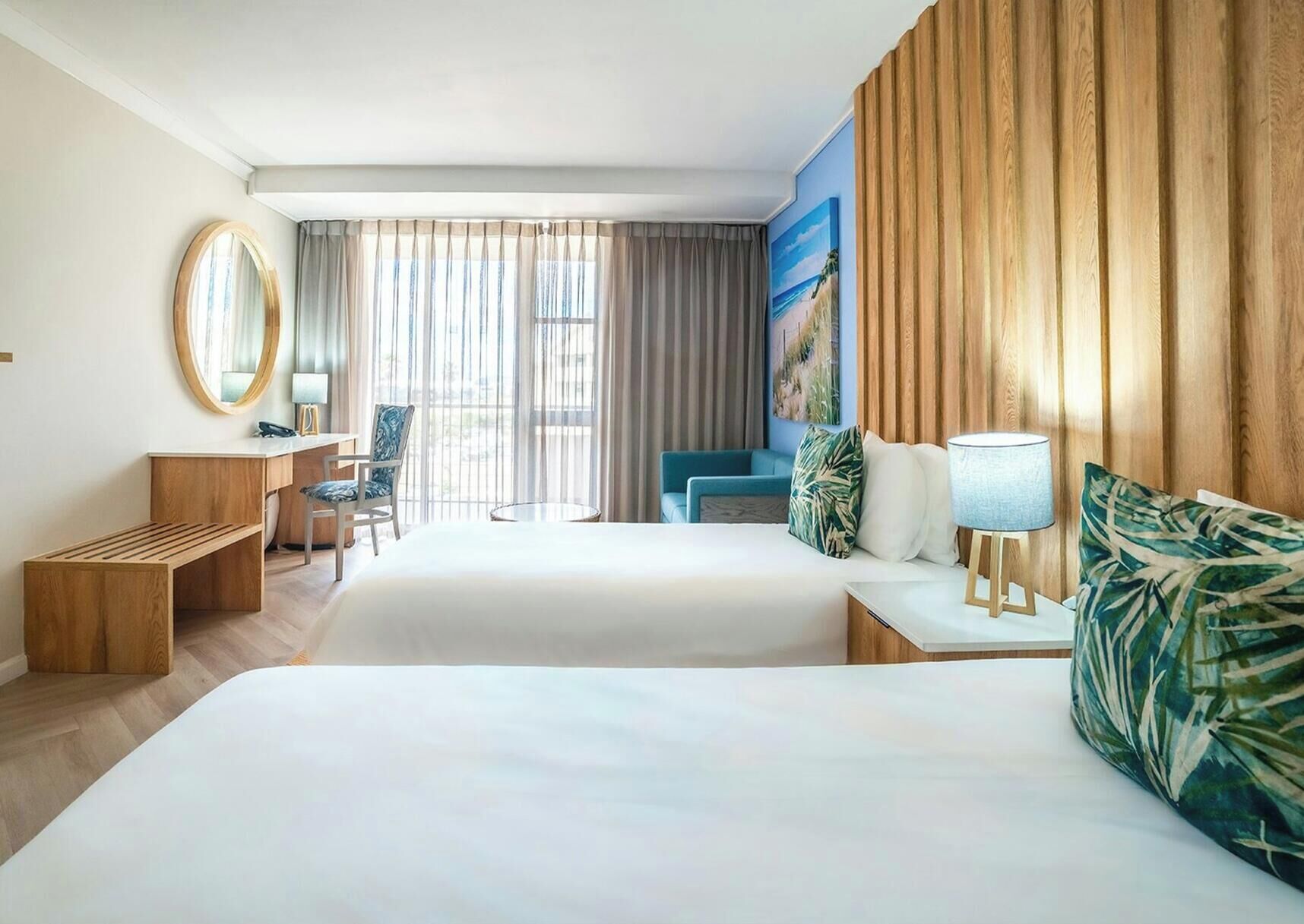 A hotel room with two beds, a desk, a chair, a round mirror, and a large window with curtains.