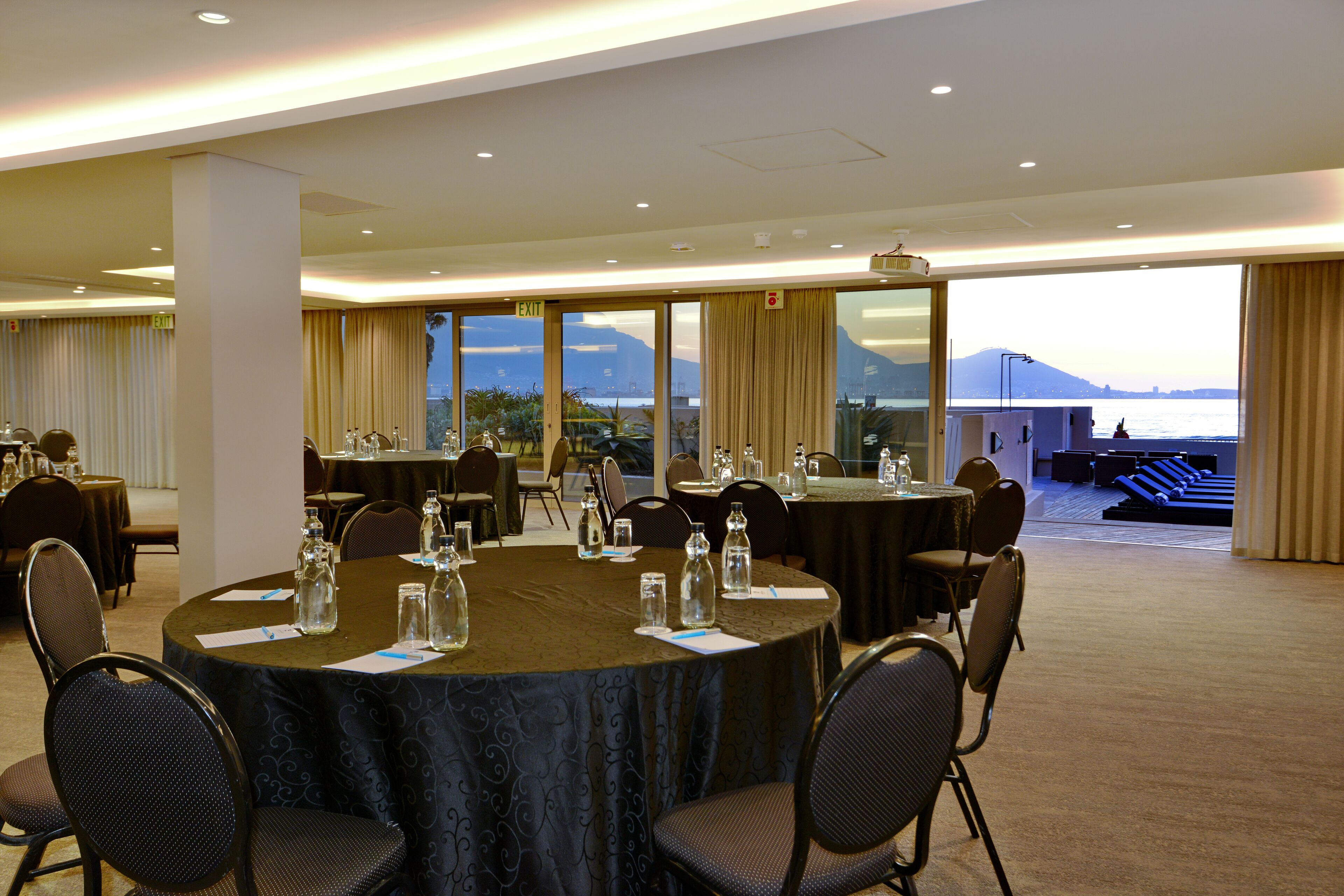 Ein Konferenzraum mit großem Tisch, Stühlen und Blick auf ein Gewässer und Berge.