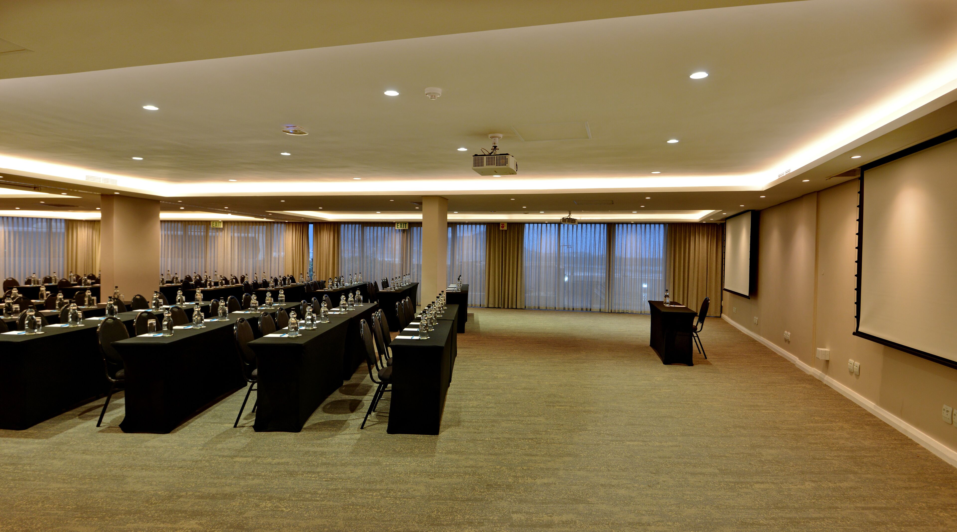 A conference room with long tables, chairs, a projector screen, and a large screen on the wall.