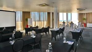 Un salón de conferencias con mesas, sillas, una pantalla de proyector y vistas a la playa.