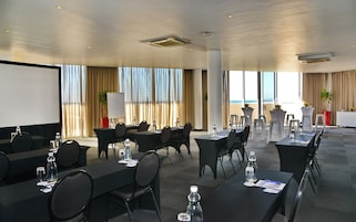 Uma sala de conferências com mesas, cadeiras, uma tela de projetor e vista para a praia.