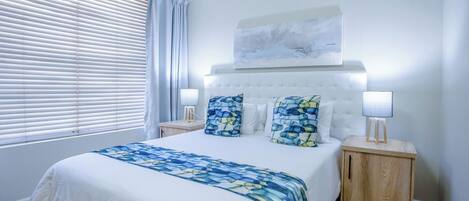 A hotel room with a large bed, two bedside tables with lamps, a window with blinds, and a painting on the wall.