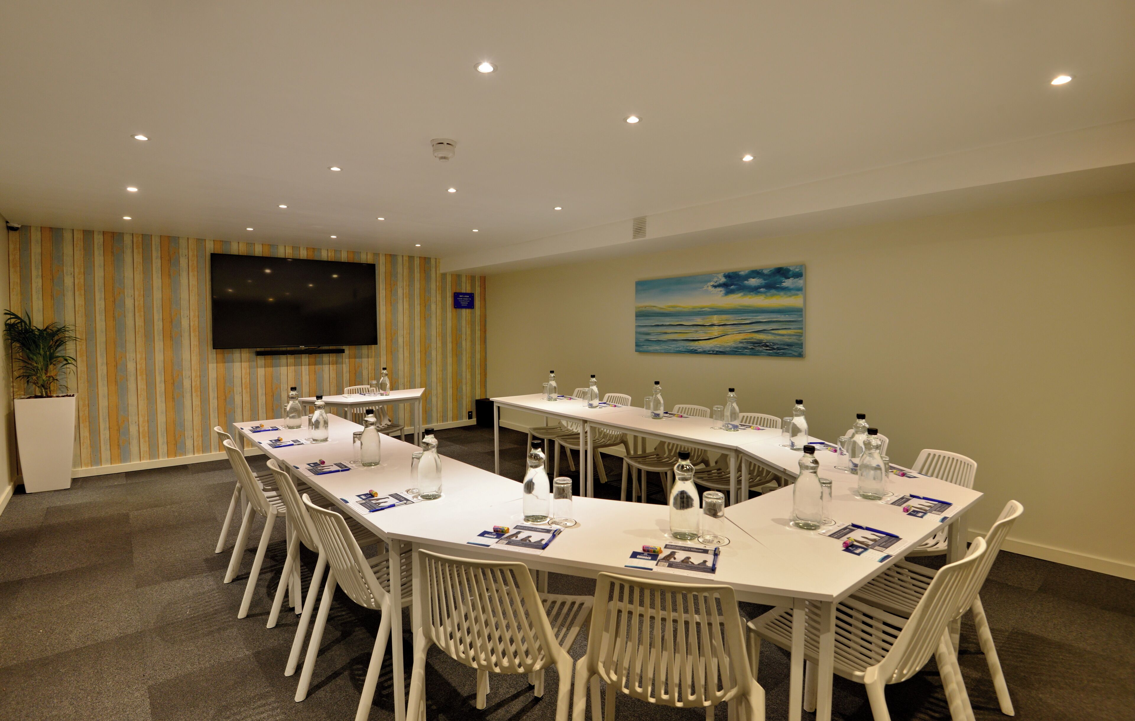 A conference room with a long table, white chairs, a flat-screen TV, and a painting on the wall.