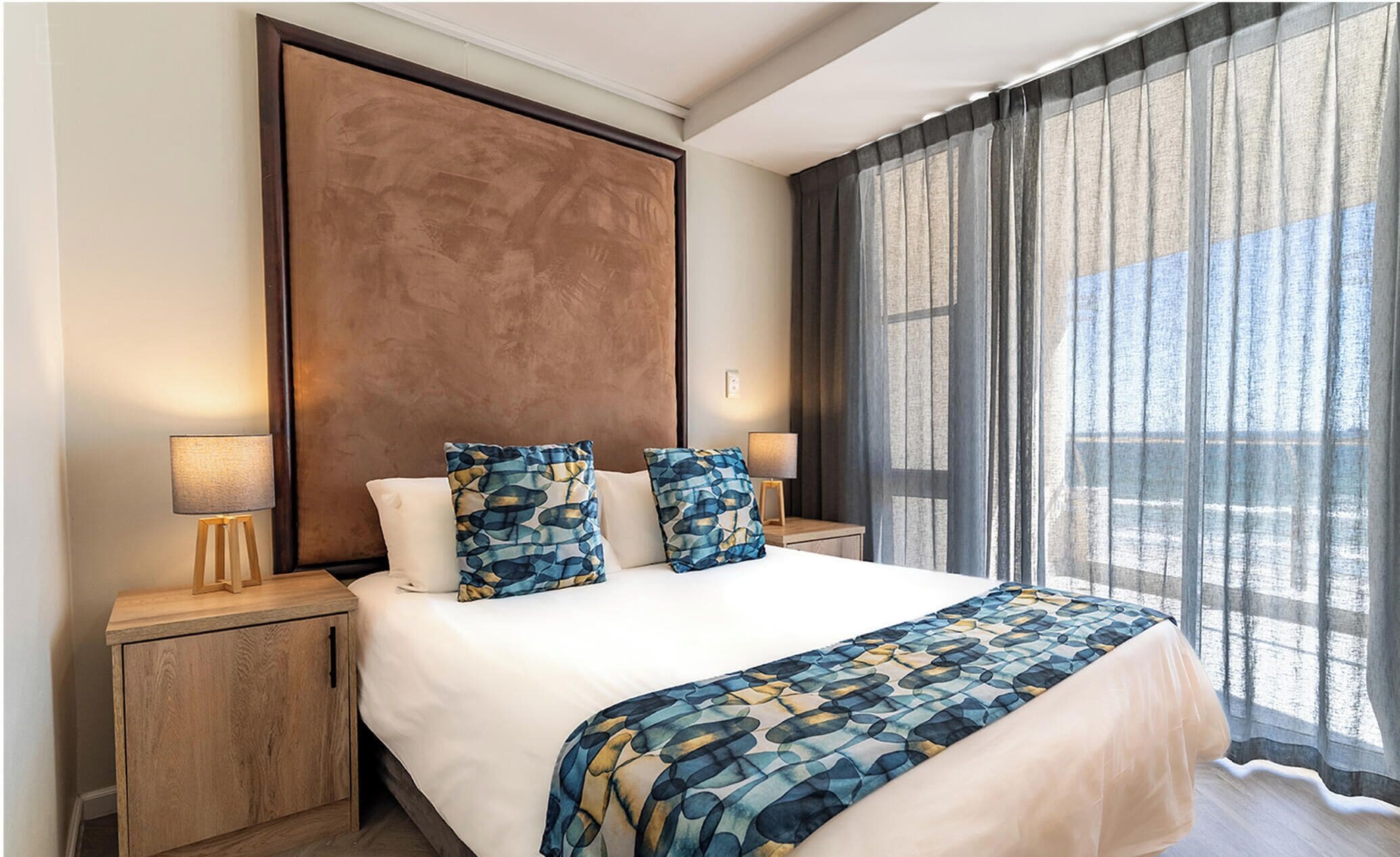 A hotel room with a large bed, two bedside lamps, a wooden nightstand, a patterned throw blanket, and sheer curtains.