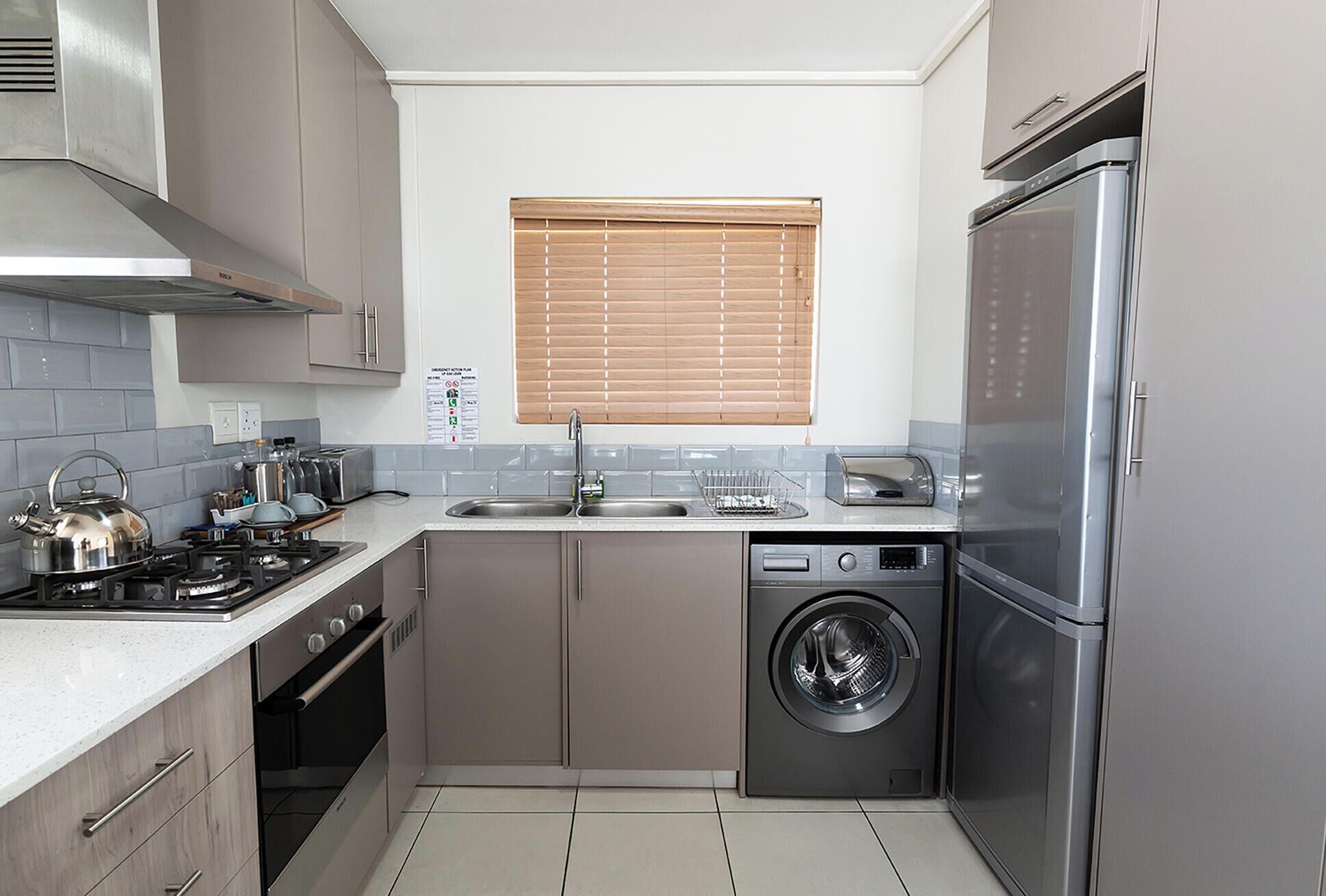 Cocina compacta con refrigerador de acero inoxidable, horno integrado, lavadora y ventana con persianas.