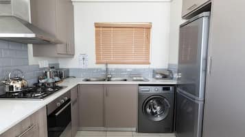 A compact kitchen with a stainless steel refrigerator, a built-in oven, a washing machine, and a window with blinds.