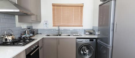 Cocina compacta con refrigerador de acero inoxidable, horno integrado, lavadora y ventana con persianas.