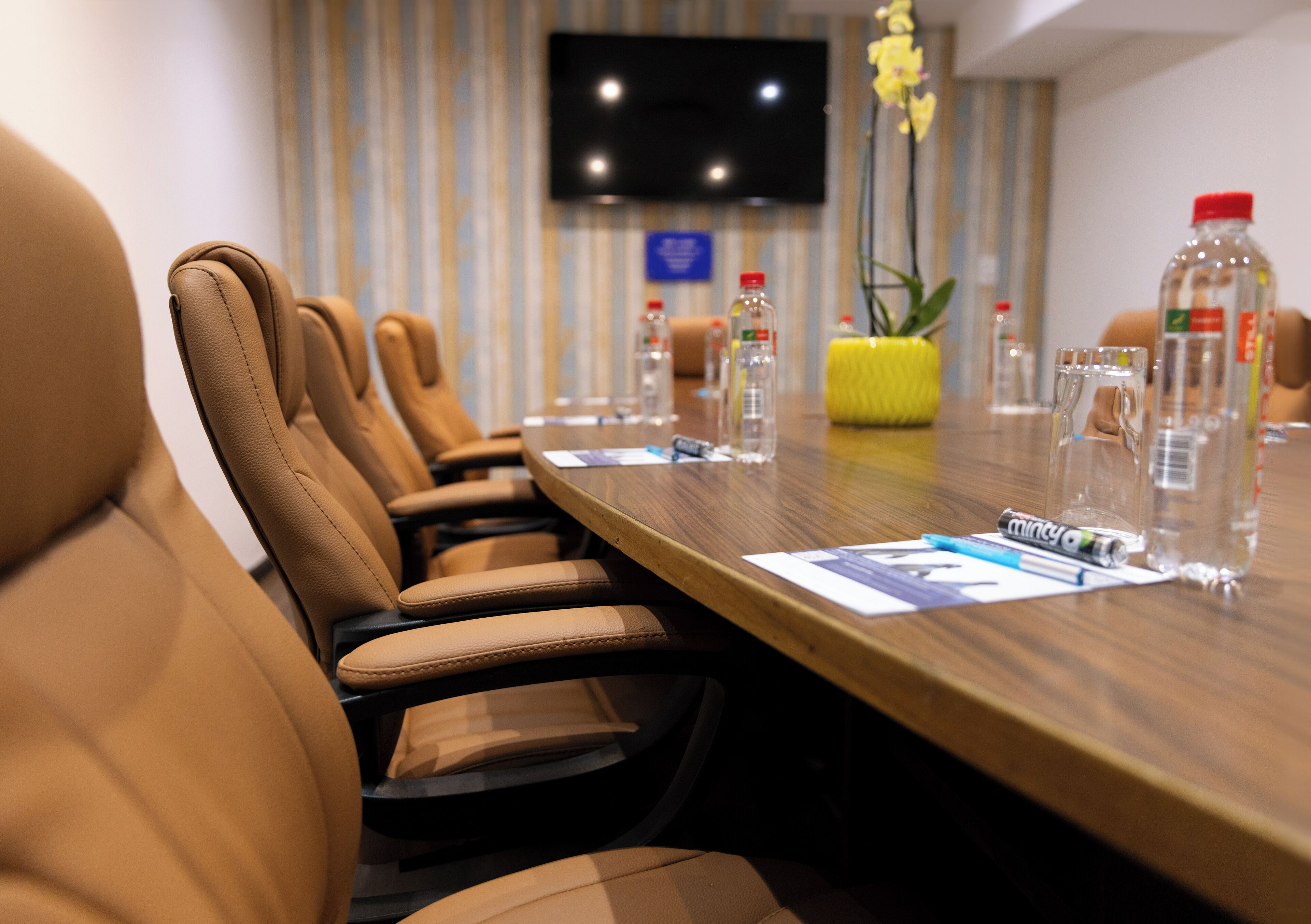 A conference room with leather chairs, a wooden table, water bottles, and a TV on the wall.