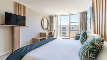 A hotel room with a large bed, a flat-screen TV, a desk with a chair, a window with curtains, and a view of buildings outside.