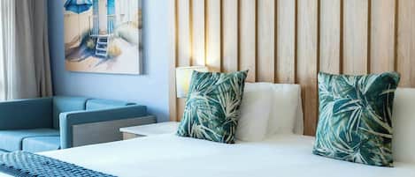 A hotel room with a bed, two pillows with a tropical leaf pattern, a blue sofa, a small table, and a painting of a beach scene.