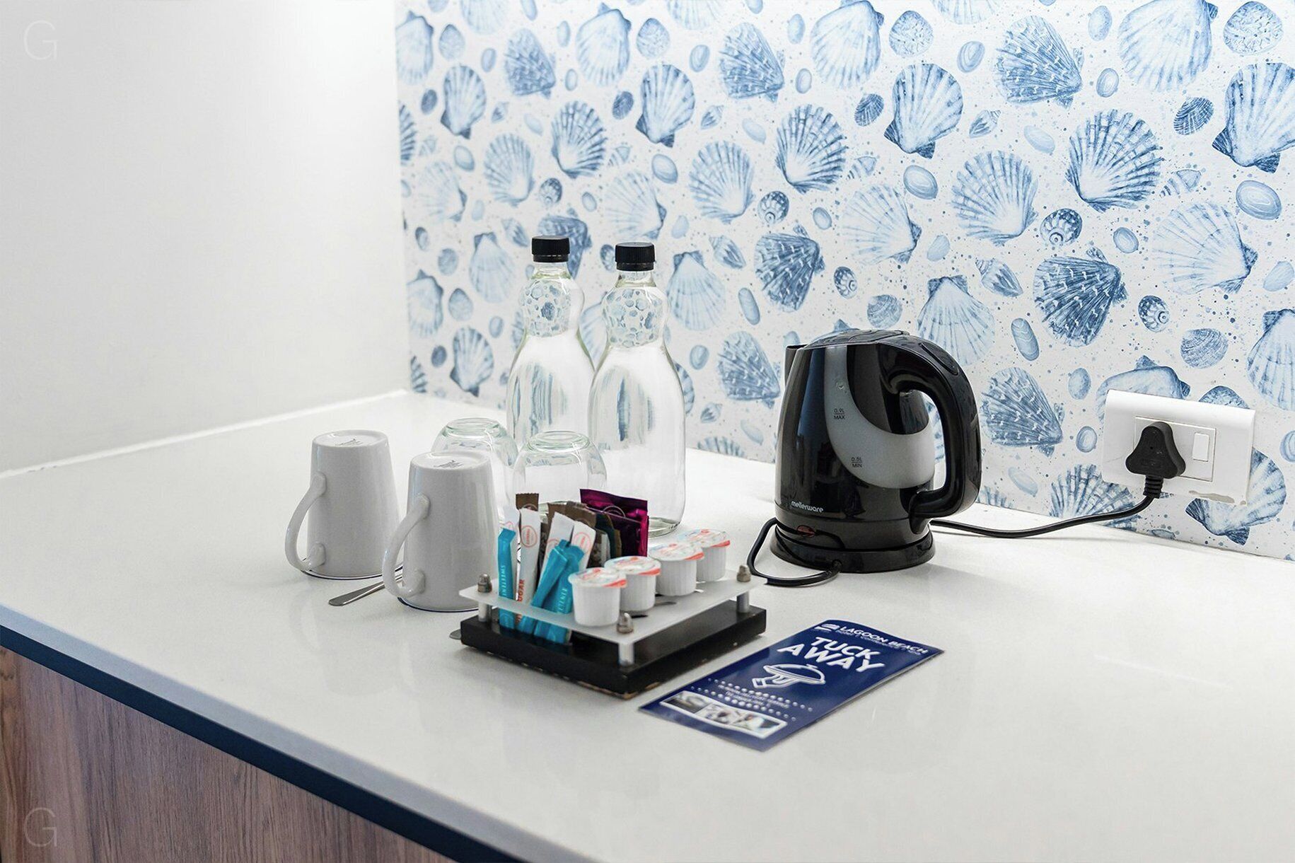 Mostrador de hotel con tetera, tazas, una botella y un perchero con artículos de aseo.