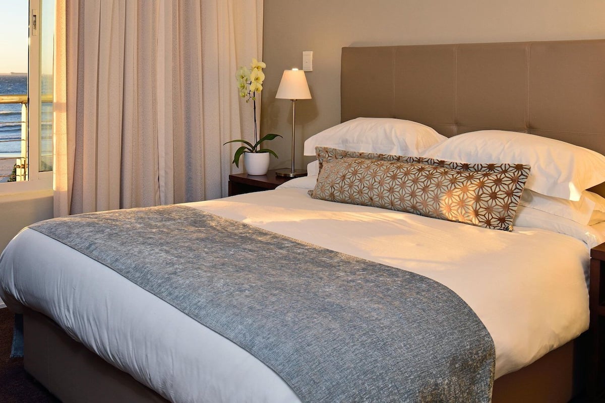 Una habitación de hotel con una cama grande, una mesita de noche, una lámpara, una planta en maceta y vistas al océano.