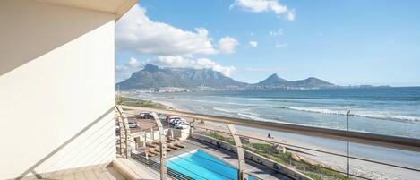 A balcony with a view of the ocean, a swimming pool, and a parking area.