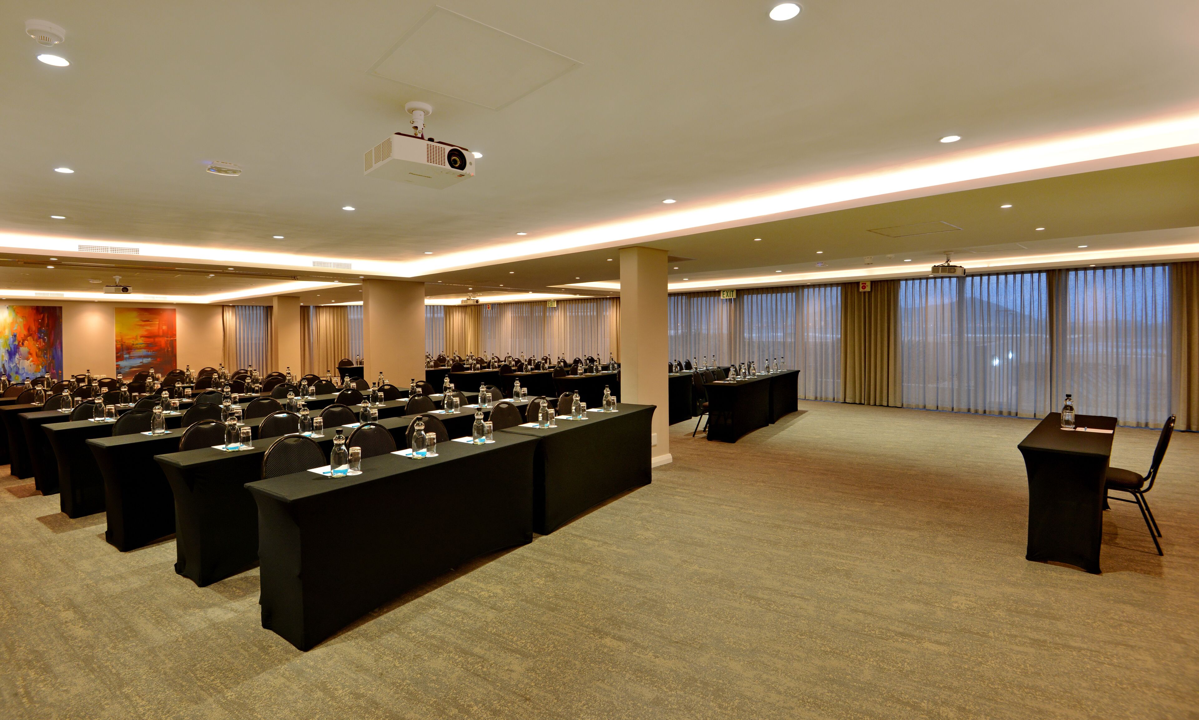 A conference room with rows of tables, chairs, and a podium, equipped with a projector and ceiling lights.