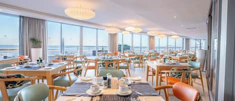 Un comedor amplio con varias mesas y sillas, ventanas grandes que ofrecen vista al mar e iluminación colgante.