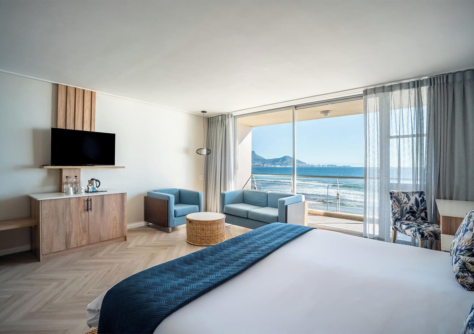 Quarto de hotel com uma cama grande, televisão, área de estar com vista para o mar e varanda com vista para a cidade.