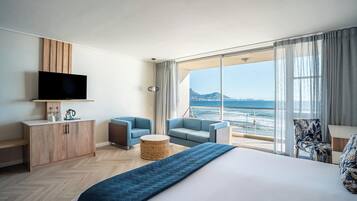 Quarto de hotel com uma cama grande, televisão, área de estar com vista para o mar e varanda com vista para a cidade.