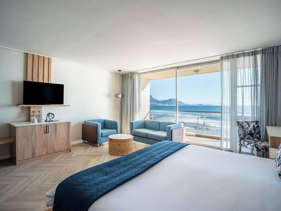 Habitación de hotel con una cama grande, televisor, zona de estar con vista al mar y balcón con vista a la ciudad.