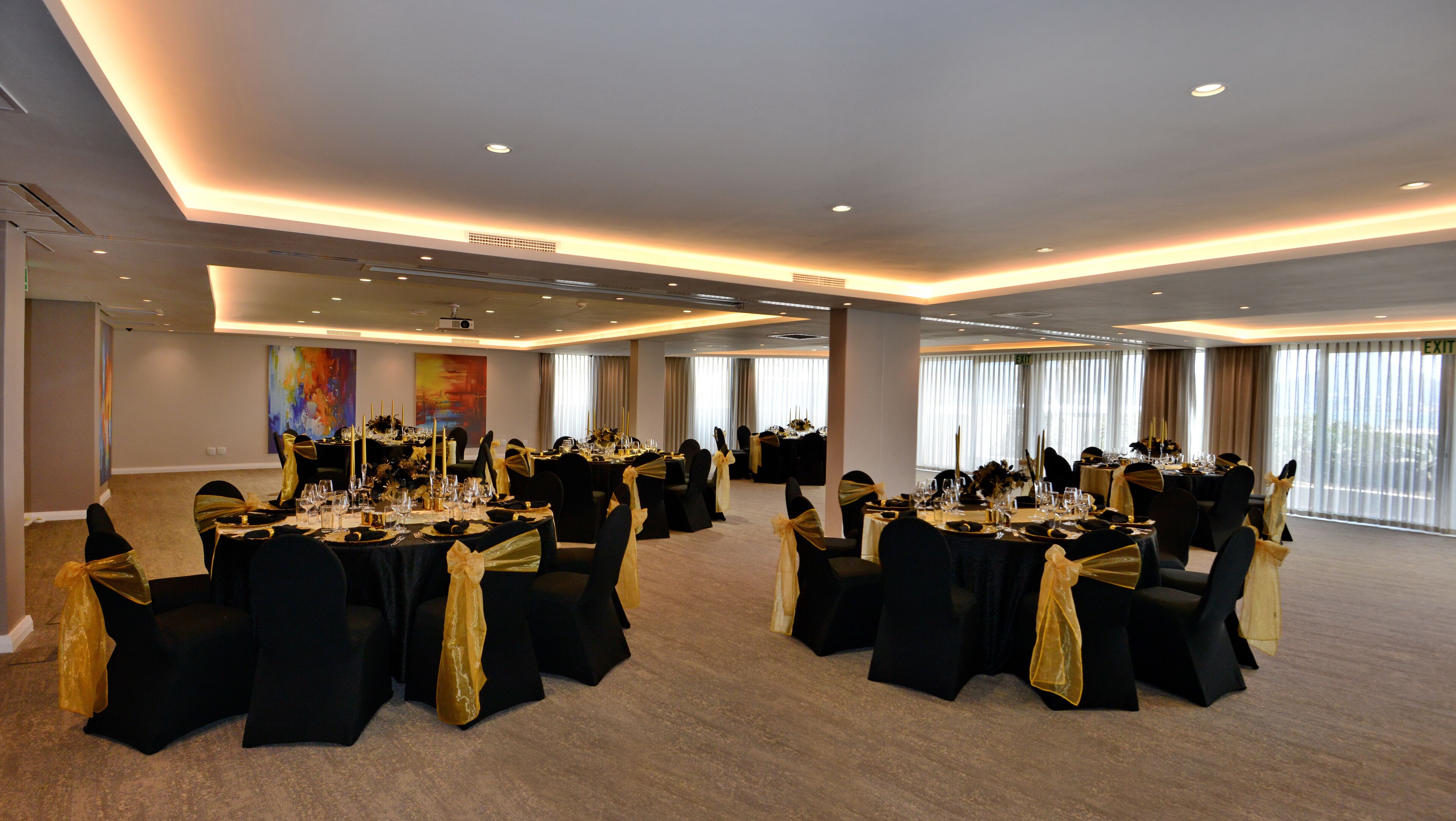 Una sala conferenze con numerosi tavoli apparecchiati per un evento formale, caratterizzata da sedie nere con nastri dorati e un soffitto moderno con illuminazione a incasso.