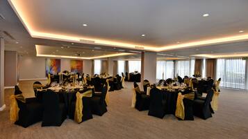 Una sala conferenze con numerosi tavoli apparecchiati per un evento formale, caratterizzata da sedie nere con nastri dorati e un soffitto moderno con illuminazione a incasso.