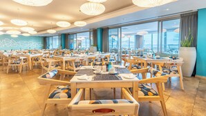 Un comedor amplio con varias mesas y sillas, ventanas grandes que ofrecen vista a la ciudad y lámparas decorativas en el techo.