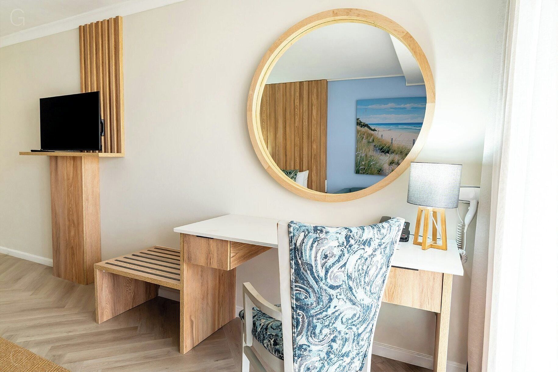 A room with a white desk, a round mirror, a television on a wooden stand, and a lamp on a small wooden table.