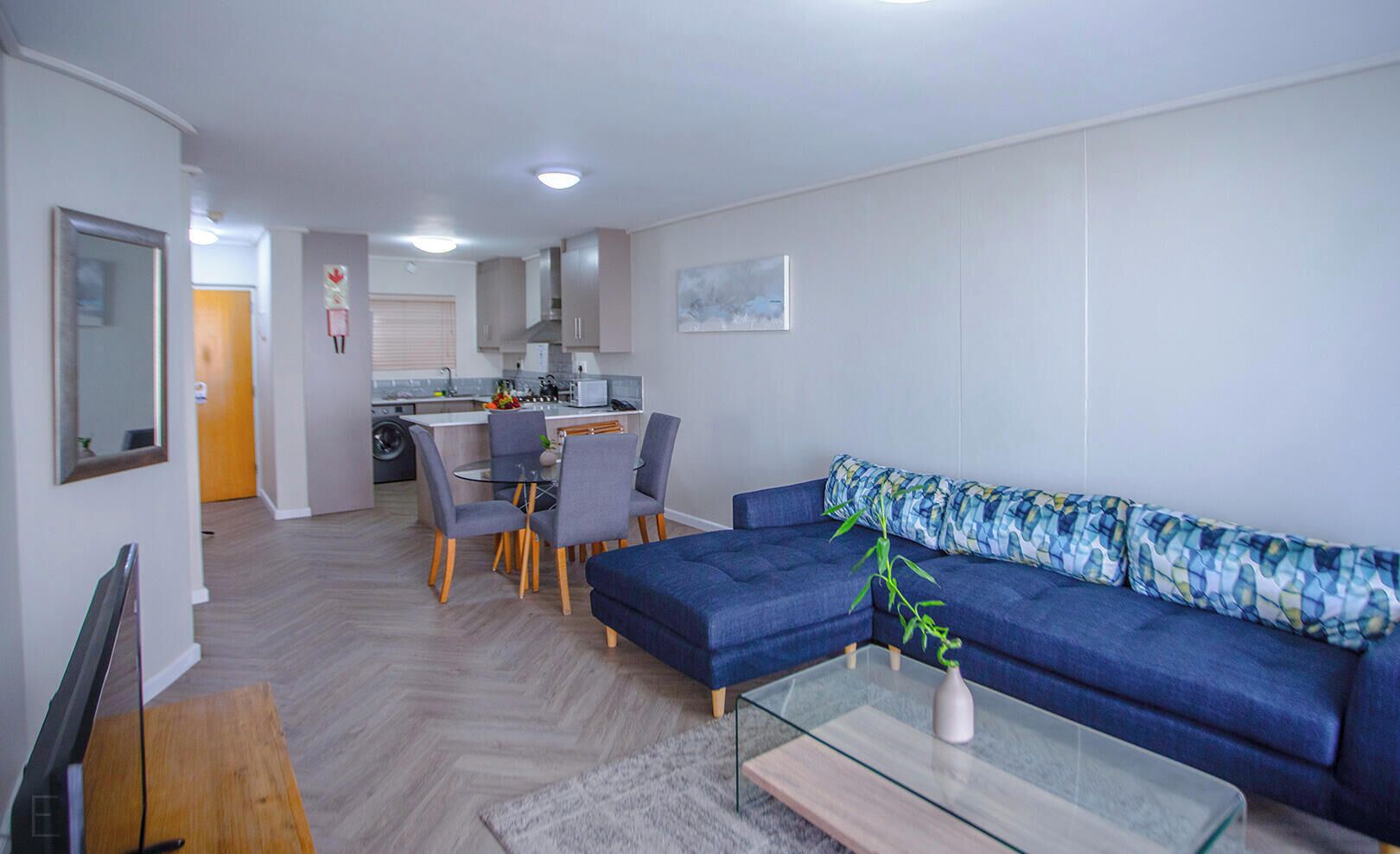A modern living room with a blue sofa, a glass coffee table, and a dining area with grey chairs.