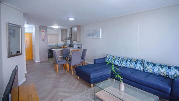 A modern living room with a blue sofa, a glass coffee table, and a dining area with grey chairs.