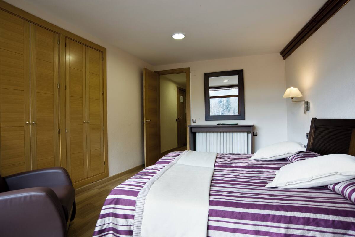 A hotel room with two beds, a wooden wardrobe, a desk with a mirror, and a lamp.
