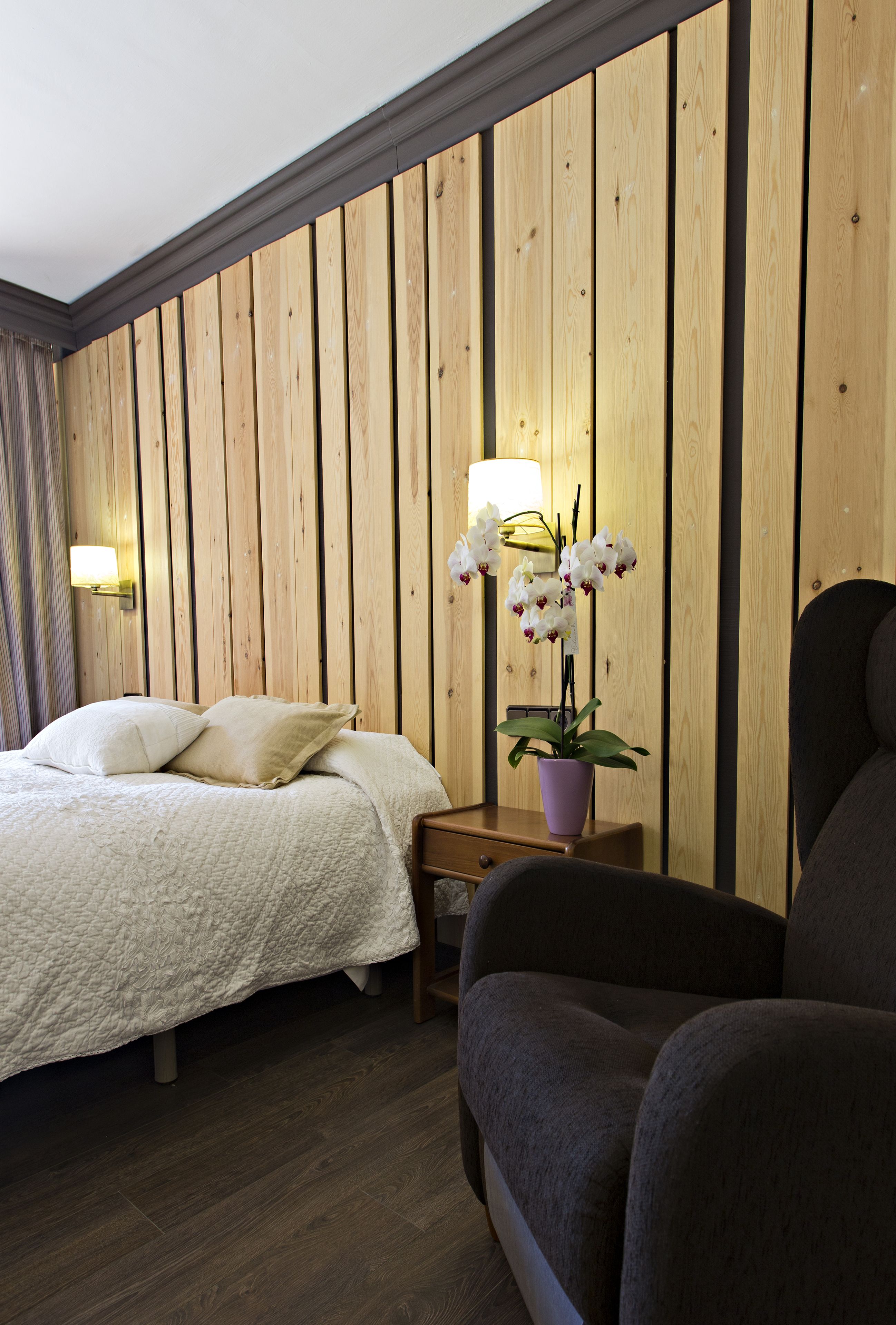 Habitación de hotel con pared de paneles de madera, cama con ropa blanca, mesita de noche con una lámpara y una orquídea en maceta, y un sillón oscuro.