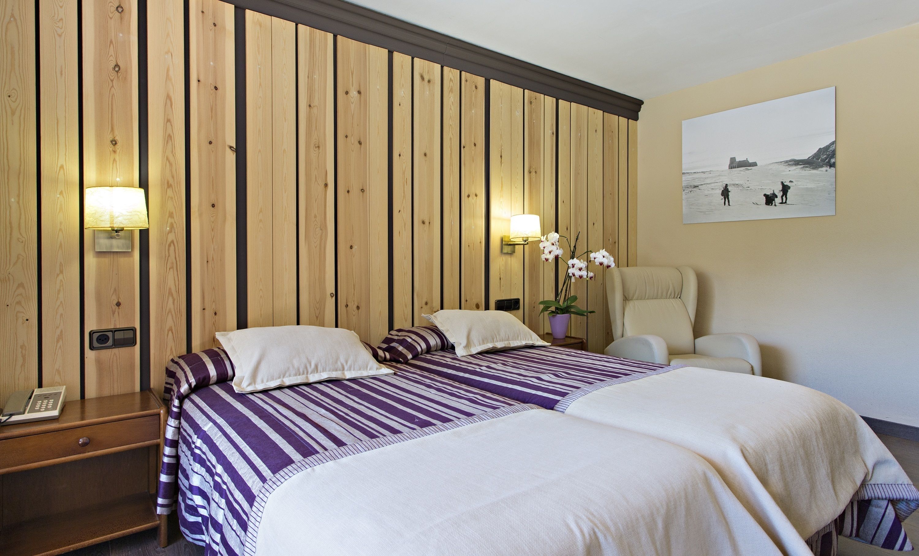 A hotel room with two beds, wooden panel walls, a painting of a beach scene, a chair, and a nightstand with a lamp.