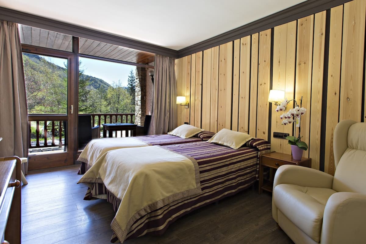 A room with two beds, a wooden wall, a balcony, a chair, and a nightstand with a lamp.