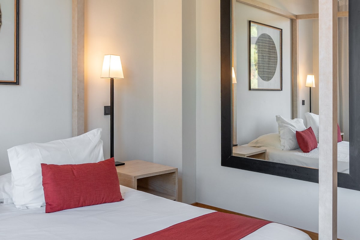 A neatly made bed with white and red bedding, a bedside lamp, and a large mirror reflecting the room.