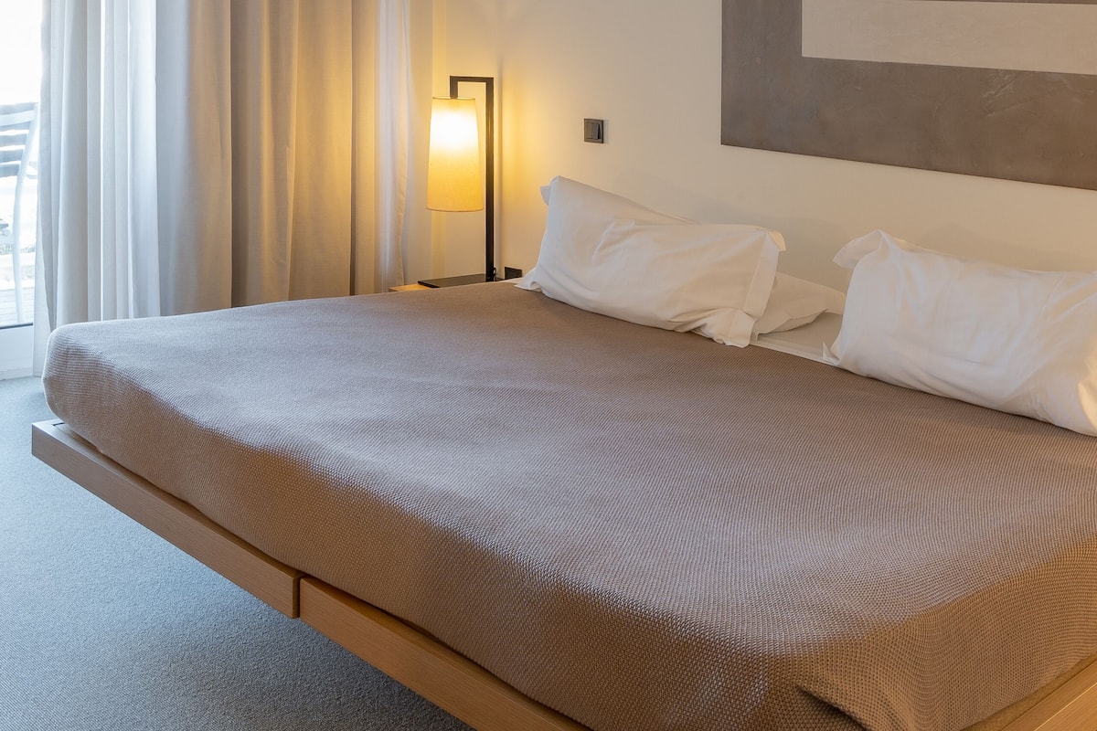 A modern hotel room with a large bed, a bedside lamp, and a view of the outdoors through a window with curtains.