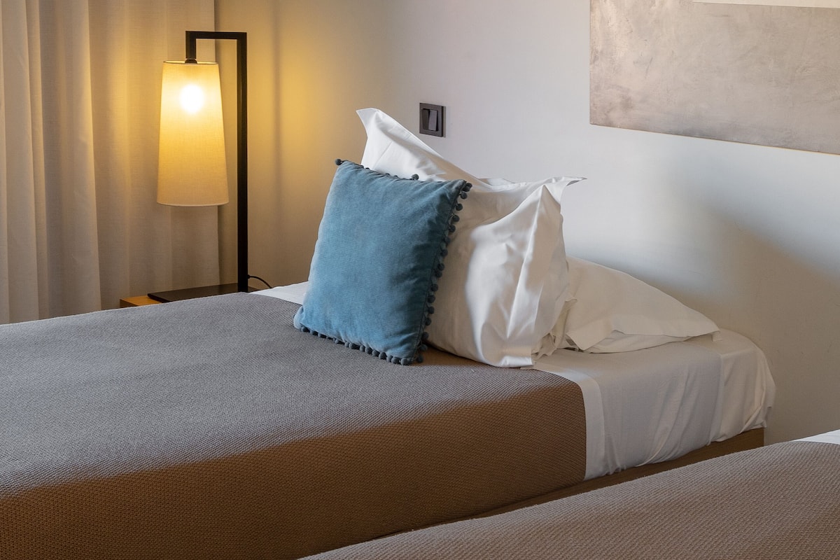 A hotel room with two beds, a bedside lamp, a wall art piece, and a window with curtains.