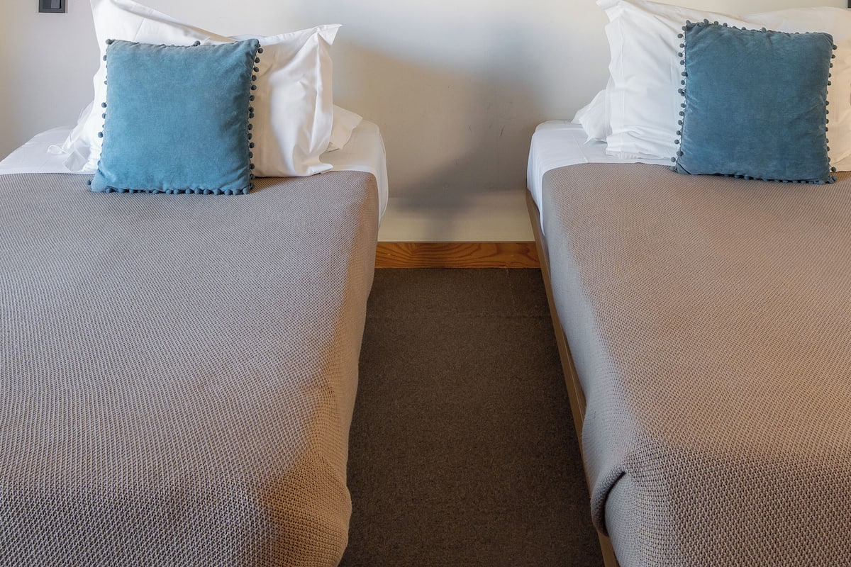 A hotel room with two beds, each with a grey blanket and a blue pillow. There is a large abstract painting on the wall above the beds.