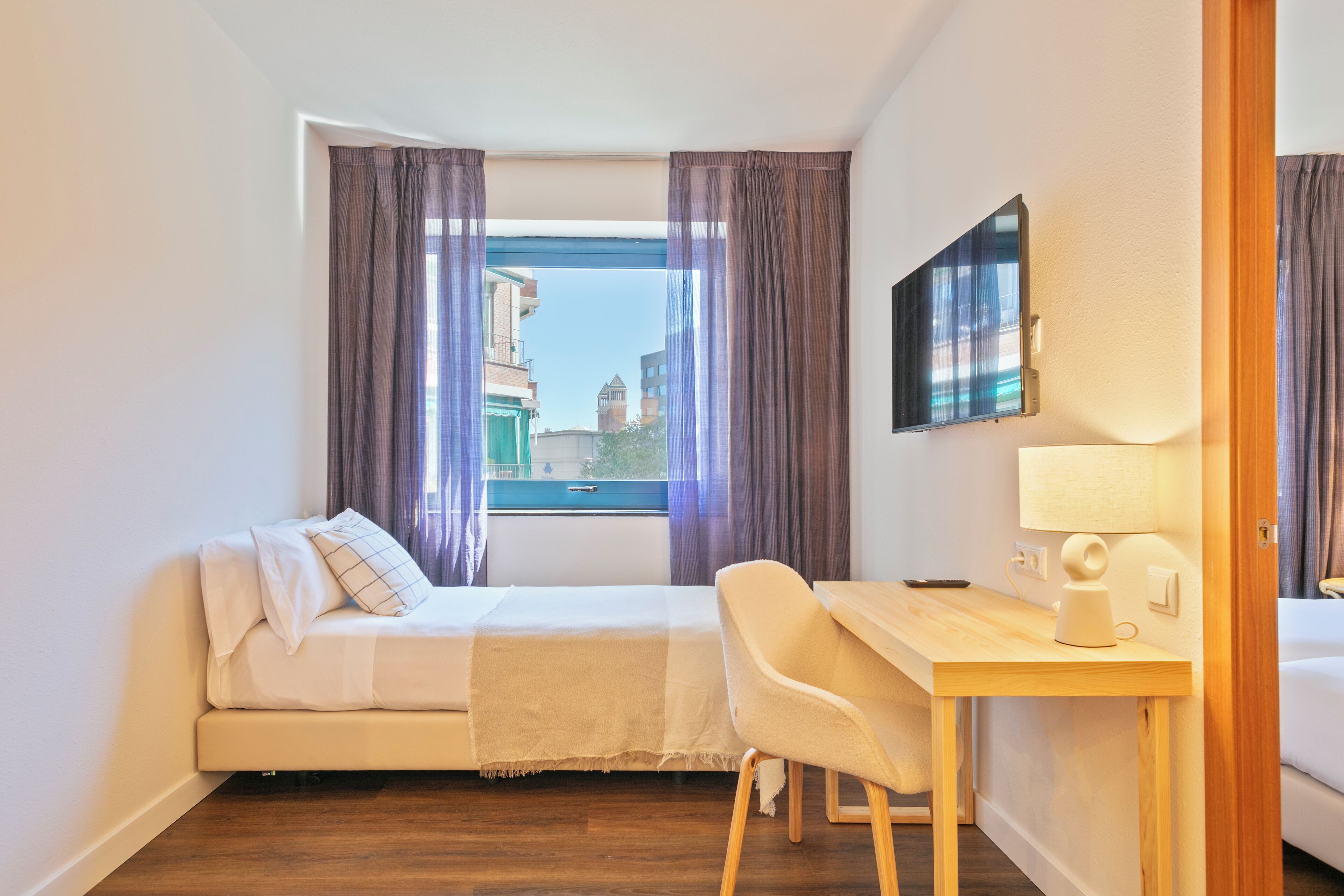 Habitación de hotel con cama, escritorio, silla, lámpara y ventana con cortinas.