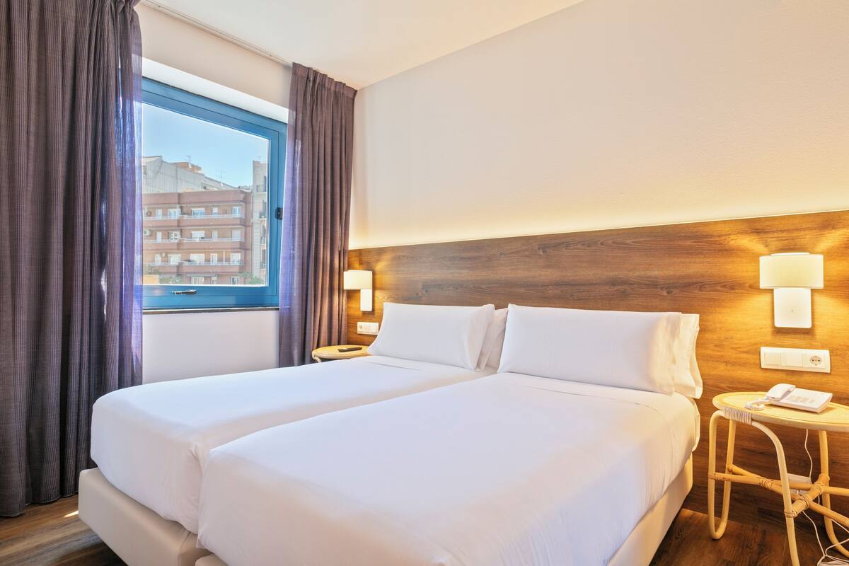 Habitación de hotel con una cama grande, una mesita de noche con lámpara, una pequeña mesa redonda con teléfono y una ventana con cortinas.