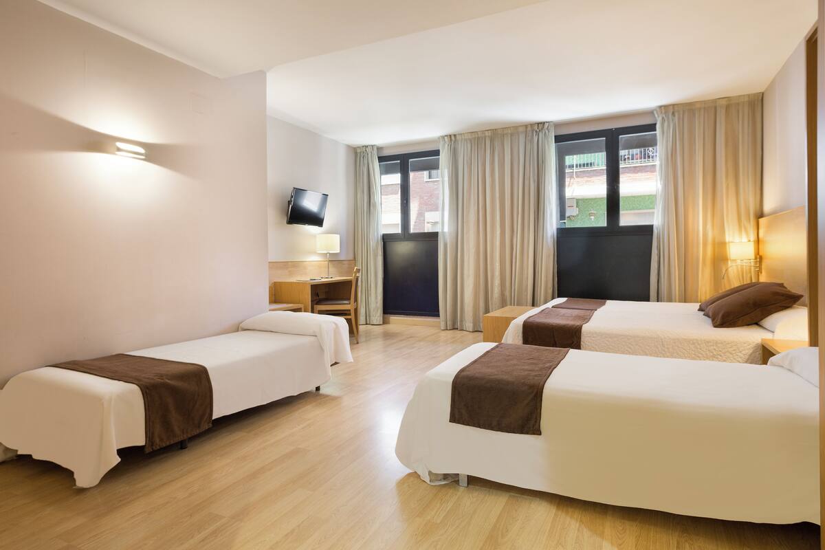 Habitación de hotel con dos camas individuales, un escritorio, una silla, un televisor y una ventana con cortinas.