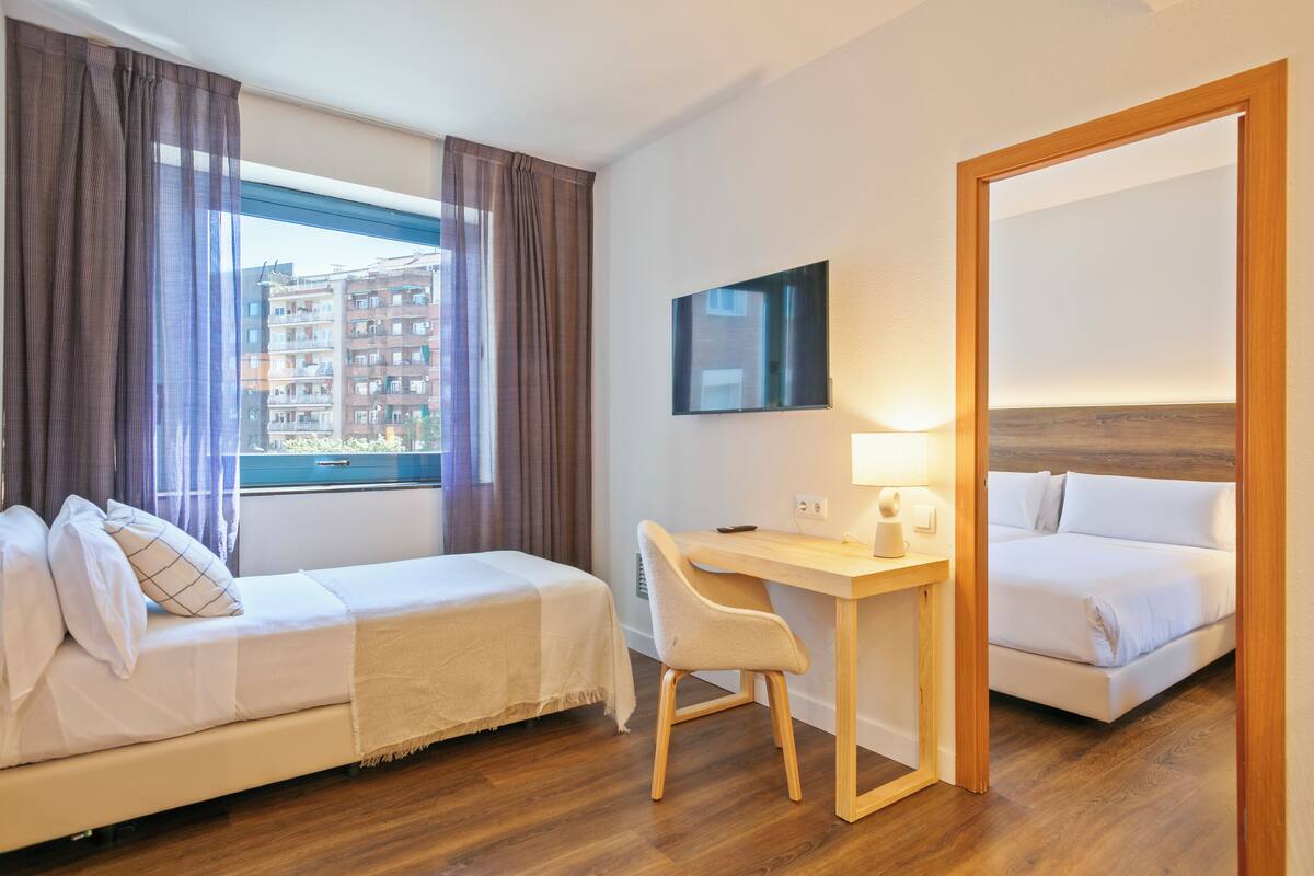 Habitación de hotel con cama, escritorio, silla, televisor y ventana con cortinas.