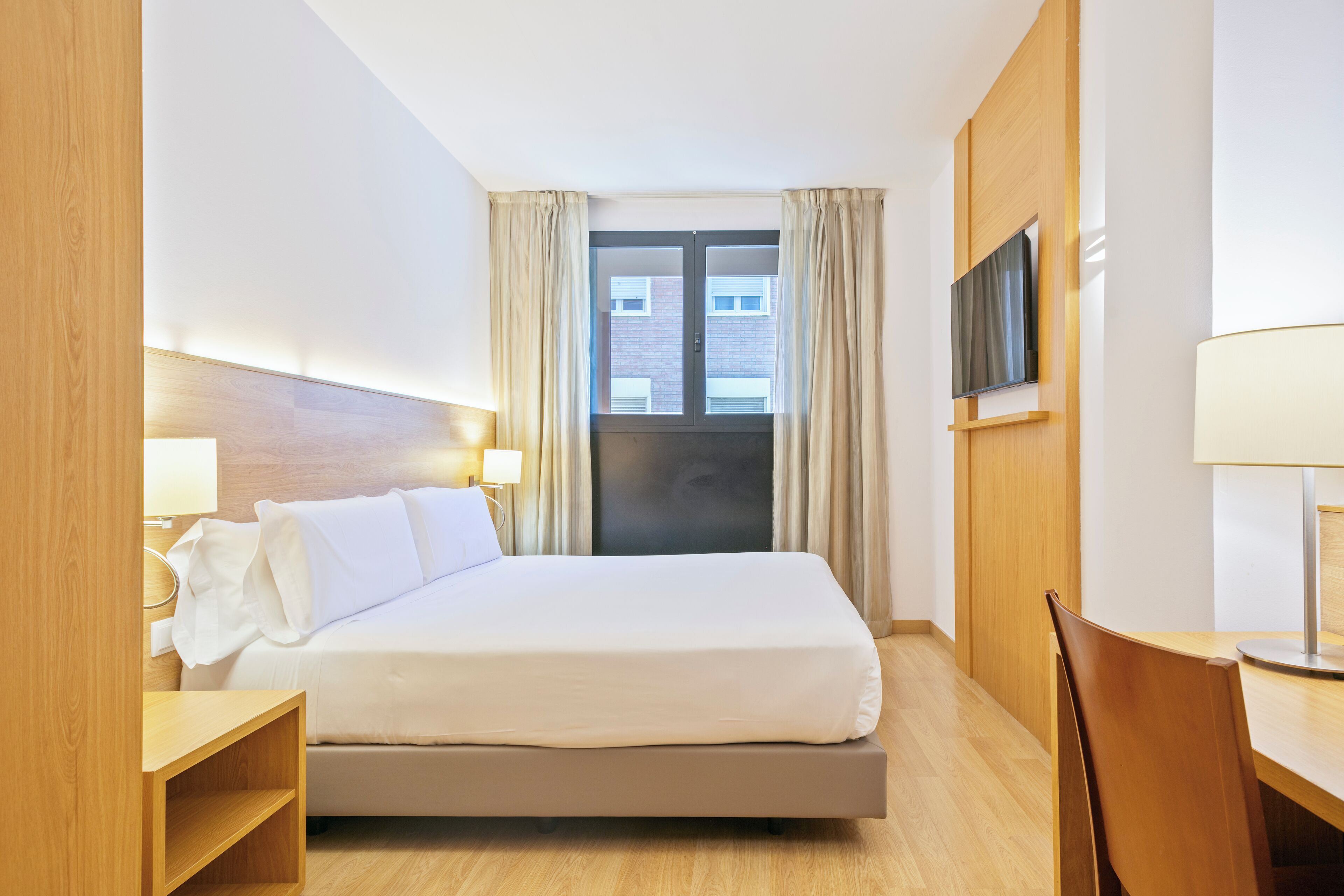 A hotel room with a bed, a desk, a chair, a TV mounted on the wall, and a window with curtains.