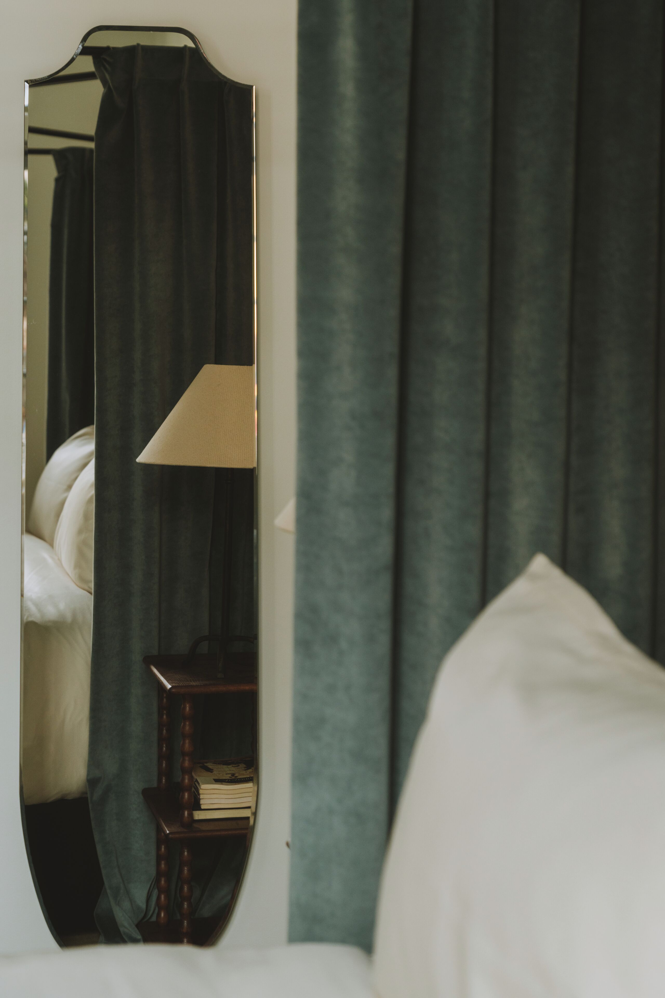 Een slaapkamer met een bed, een nachtkastje met een lamp, en een spiegel die een gordijn en een lamp reflecteert.