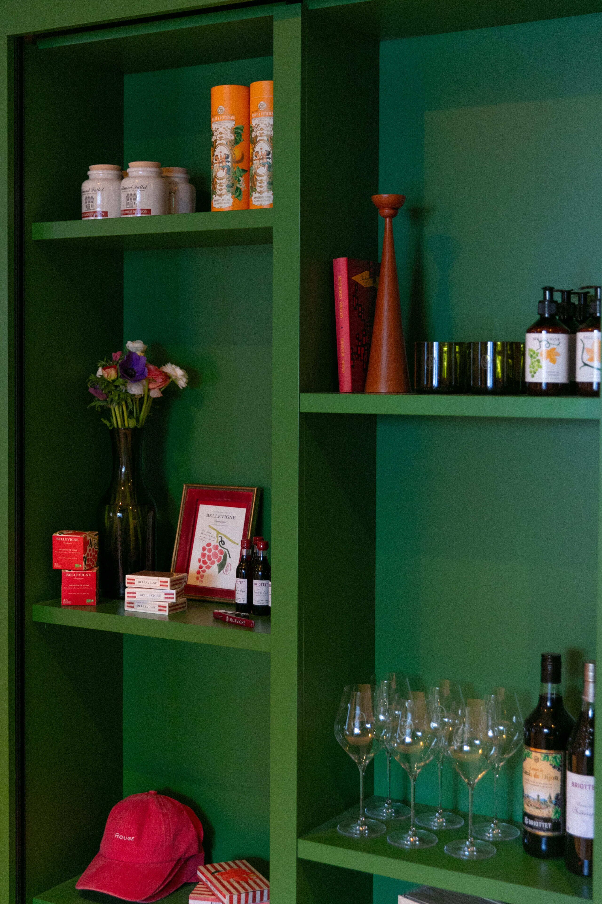 A green shelf with wine bottles, glasses, a vase with flowers, and a red cap.